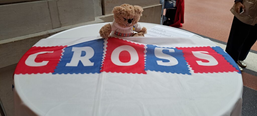 Ein Teddy mit Postcrossing Shirt sitzt auf einem runden Tisch neben einer Postcrossing Fahne