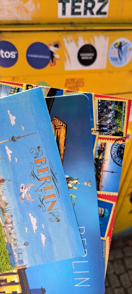3 Ansichtskarten im Fächer gehalten vor einem gelben Briefkasten
