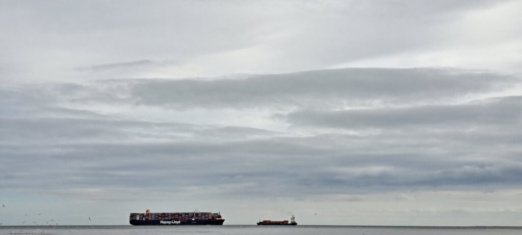 Ein Riesencontainerschiff und ein kleineres Schiff fahren am Horizont auf der Nordsee