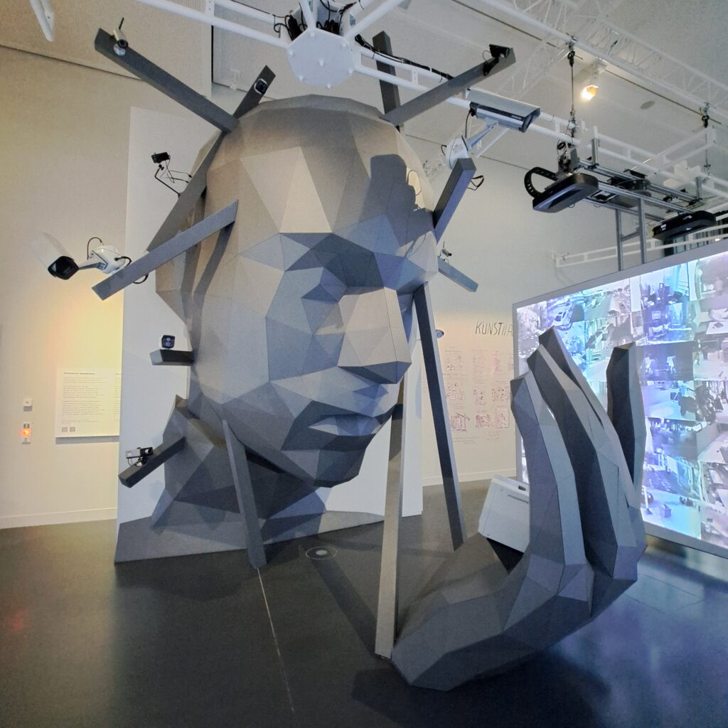 Raumgroßer Halbschädel aus grauem Kunststoff, der auf seine Hand blickt, in der ein Monitor ist und dem Überwachungskameras auf dem Kopf ragen.